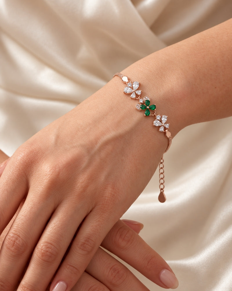 A delicate rose gold silver 925 bracelet with three floral clusters, featuring clear crystals and one green emerald, worn on a woman's wrist against a cream satin background.