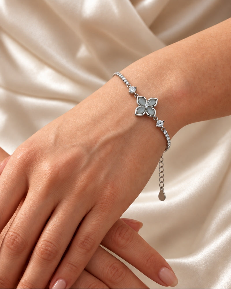 A delicate 925 sterling silver bracelet with a four-petal flower motif and small clear stones, shown worn on a person's wrist against a soft cream satin background.