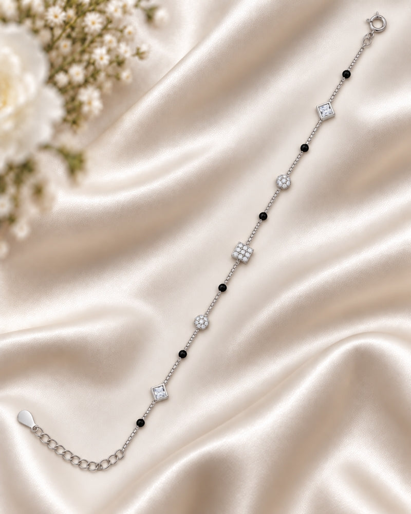 A delicate silver chain bracelet with alternating black beads and clear crystal geometric charms, displayed on a draped champagne satin fabric with white flowers in the corner.