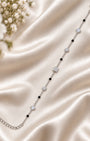 A delicate silver chain bracelet with alternating black beads and clear crystal geometric charms, displayed on a draped champagne satin fabric with white flowers in the corner.