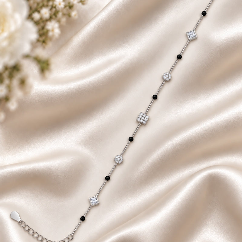 A delicate silver chain bracelet with alternating black beads and clear crystal geometric charms, displayed on a draped champagne satin fabric with white flowers in the corner.