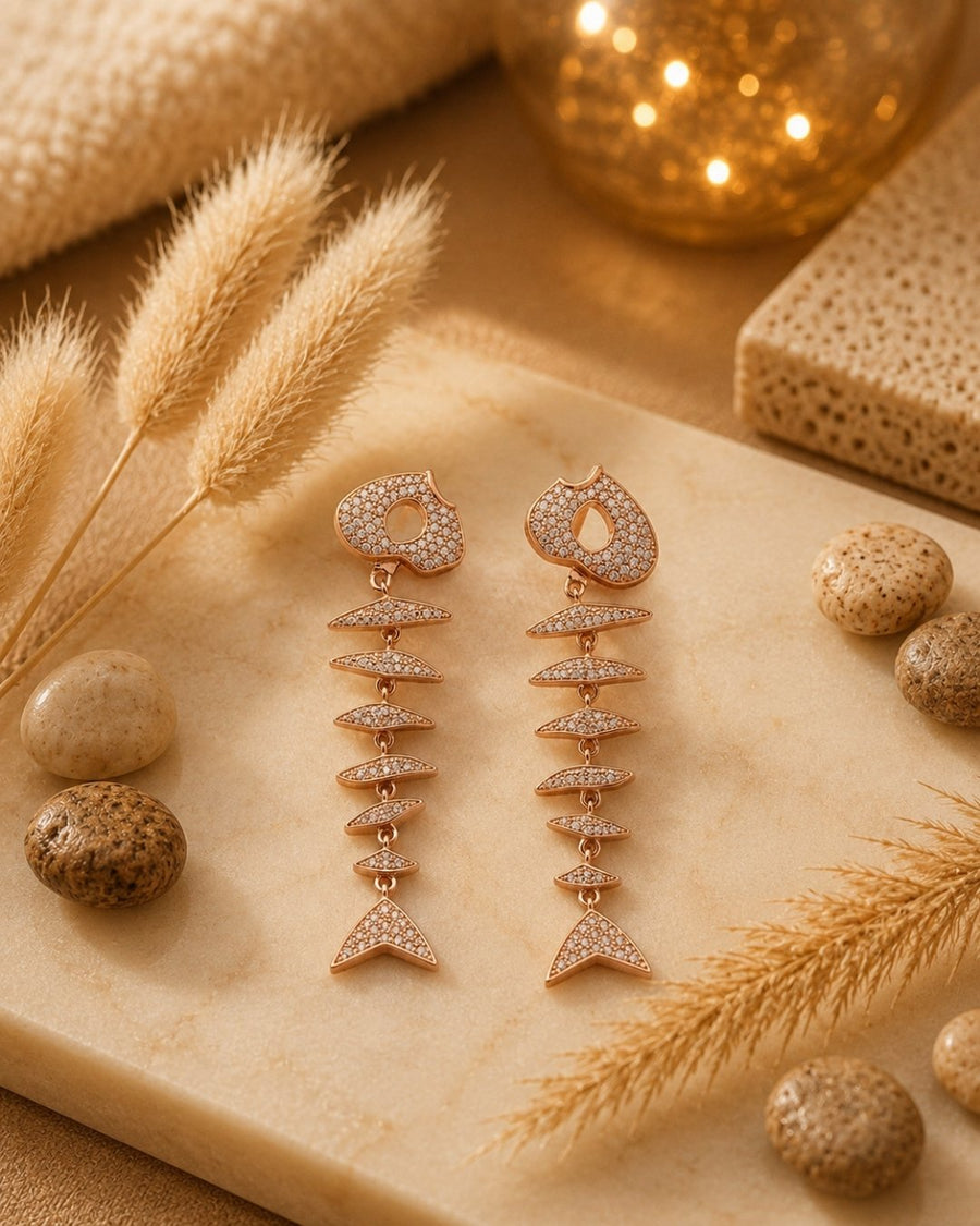 A pair of dangling fish-scale silver 925 earrings in rose gold with pavé-set clear stones, styled on a beige stone slab with dried grass, decorative stones, and warm ambient lighting.