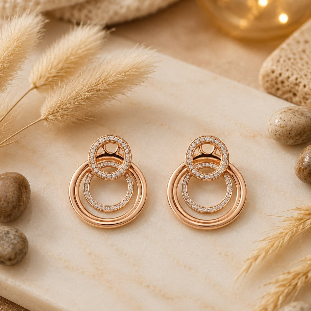 A pair of rose gold double-ring silver 925 drop earrings with small clear pavé accents, displayed on a cream marble slab with dried grass, stones, and warm bokeh lights in the background.