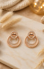 A pair of rose gold double-ring silver 925 drop earrings with small clear pavé accents, displayed on a cream marble slab with dried grass, stones, and warm bokeh lights in the background.