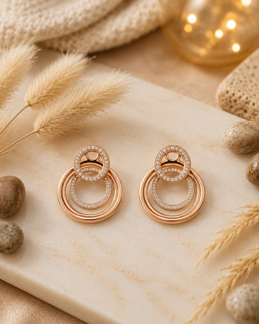 A pair of rose gold double-ring silver 925 drop earrings with small clear pavé accents, displayed on a cream marble slab with dried grass, stones, and warm bokeh lights in the background.