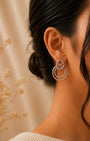 A close-up side profile of a woman wearing a pair of silver 925 double-ring hoop earrings adorned with small clear stones, set against a warm beige background with dried floral decor.