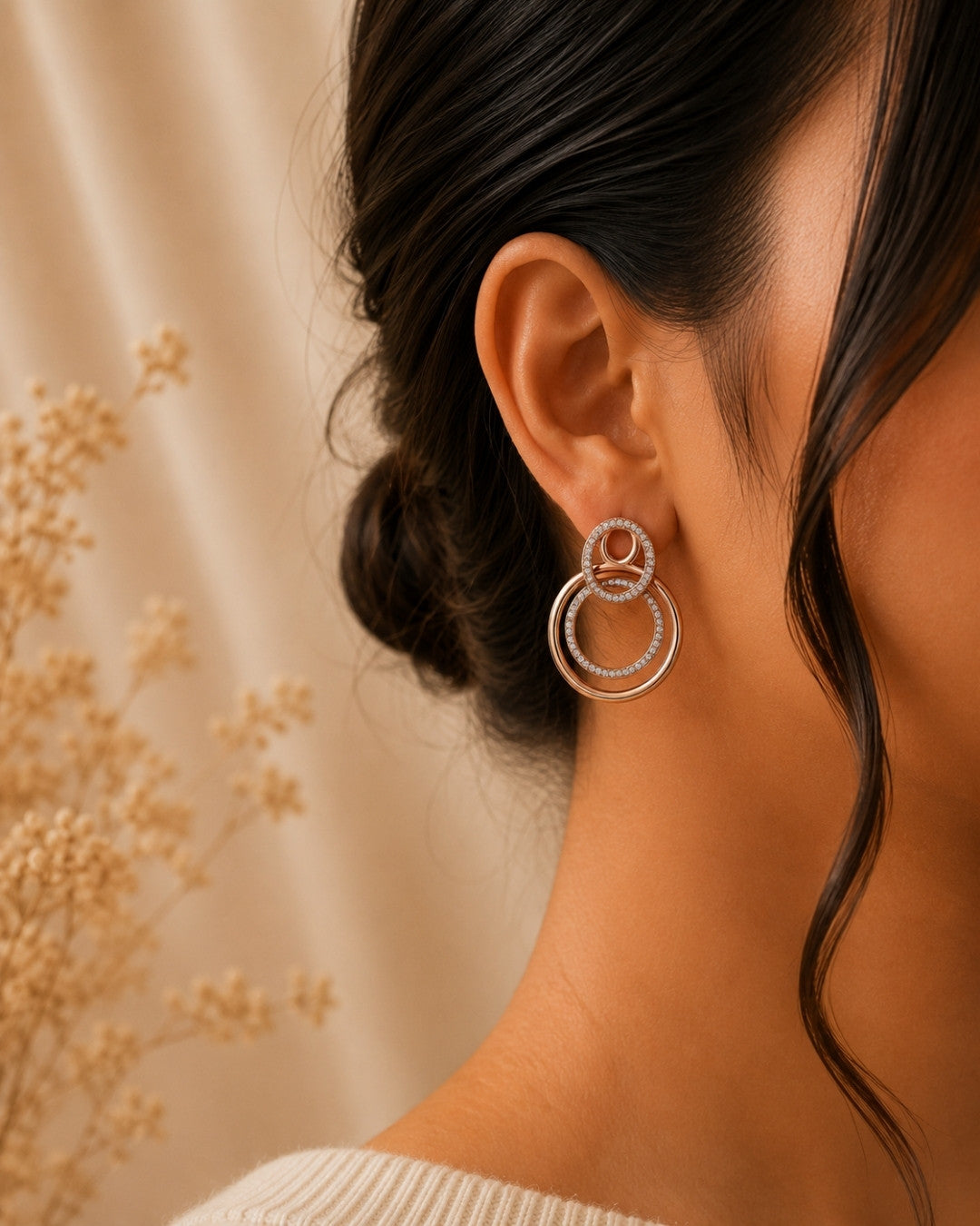 A close-up side profile of a woman wearing a pair of silver 925 double-ring hoop earrings adorned with small clear stones, set against a warm beige background with dried floral decor.