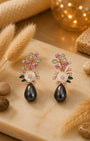 A pair of ornate floral drop silver 925 earrings displayed on a beige stone slab with dried grass, small stones, and a warm glowing decorative sphere in the background. The earrings feature pink and green gemstone floral tops, white flower centers, and large dark teardrop pearls hanging below.