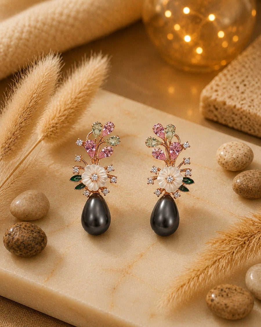 A pair of ornate floral drop silver 925 earrings displayed on a beige stone slab with dried grass, small stones, and a warm glowing decorative sphere in the background. The earrings feature pink and green gemstone floral tops, white flower centers, and large dark teardrop pearls hanging below.