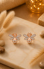 A pair of rose gold silver 925 bee stud earrings with pavé-set clear stones, displayed on a beige marble slab surrounded by dried grass, decorative stones, and a warm glowing background.