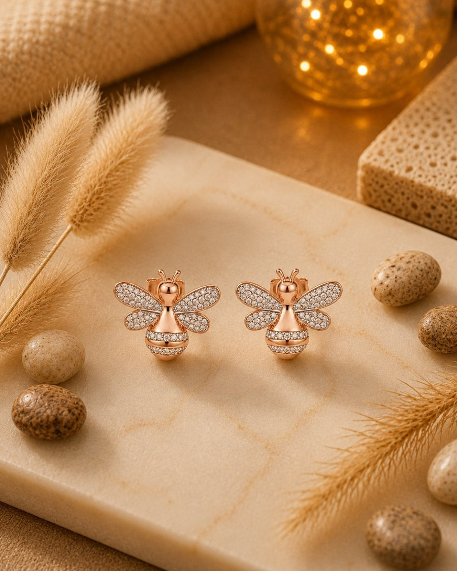 A pair of rose gold silver 925 bee stud earrings with pavé-set clear stones, displayed on a beige marble slab surrounded by dried grass, decorative stones, and a warm glowing background.