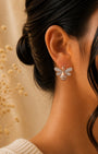 A close-up of a woman's ear wearing a delicate silver 925 bee-shaped stud earring with sparkling clear stones and a gold-toned center, against a warm beige background with dried floral decor.
