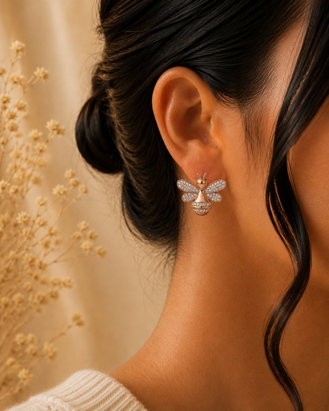 A close-up of a woman's ear wearing a delicate silver 925 bee-shaped stud earring with sparkling clear stones and a gold-toned center, against a warm beige background with dried floral decor.